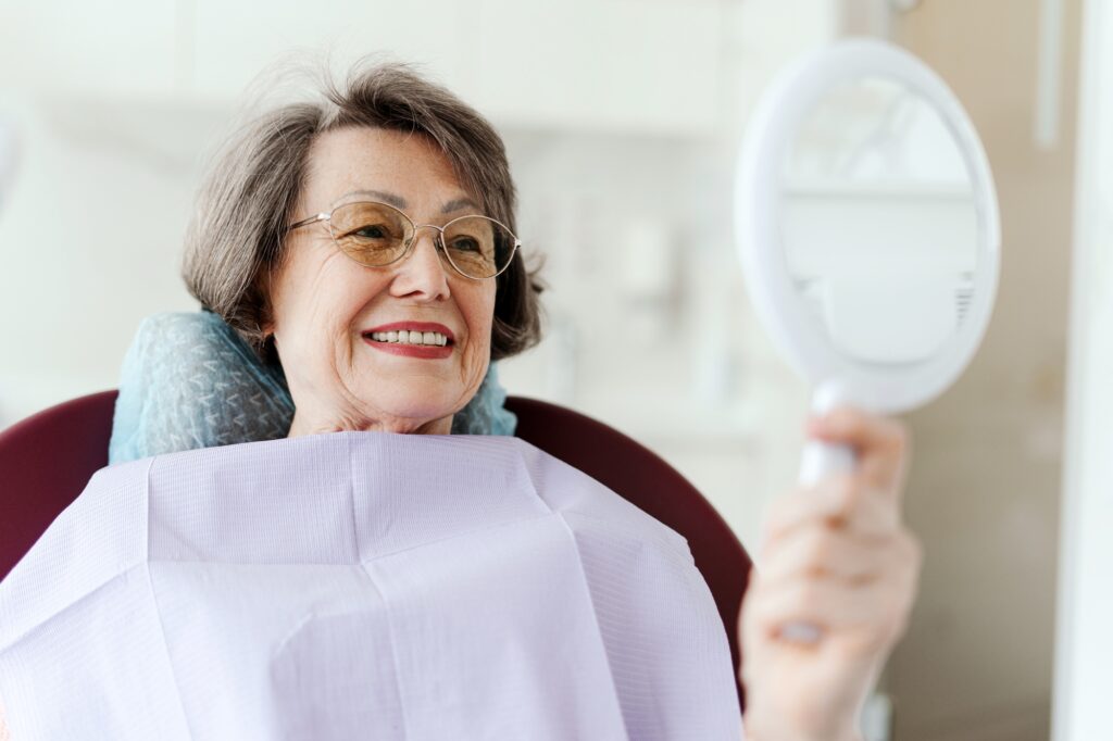 Smiling senior female patient holding mirror looking at teeth while sitting in dental chair